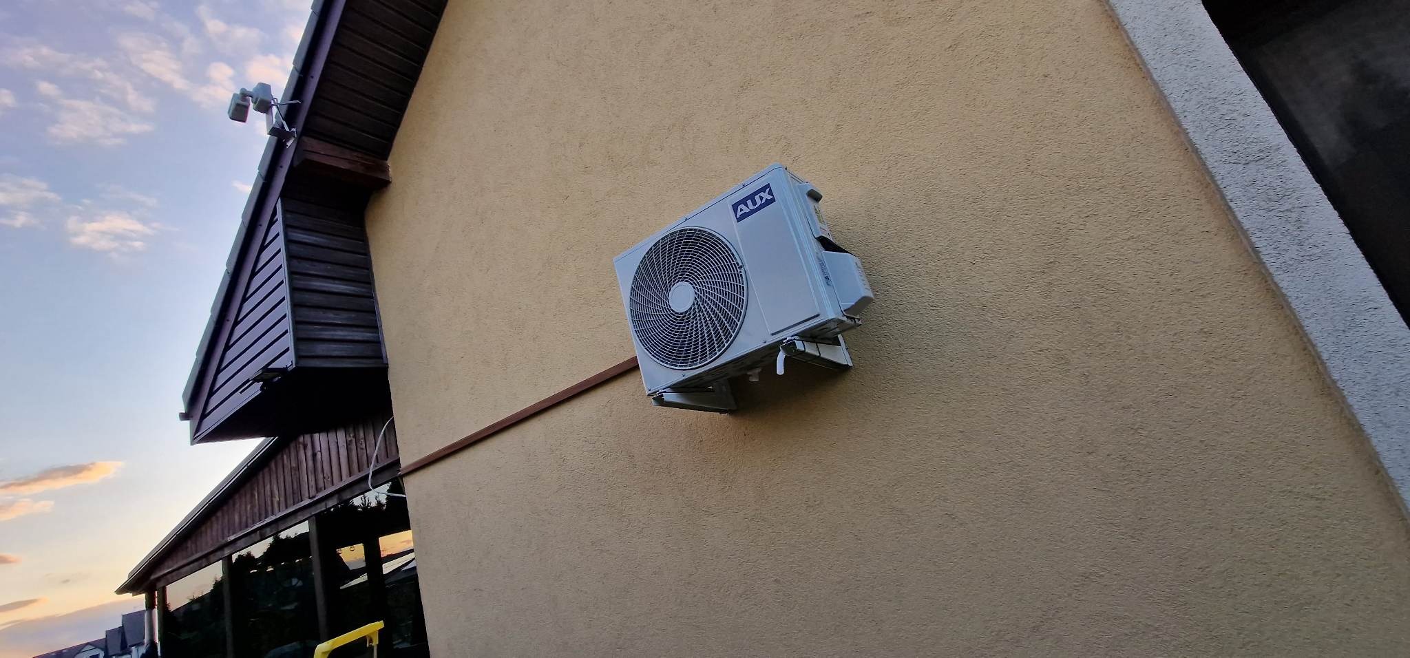 Zewnętrzna jednostka klimatyzacji marki AUX zamontowana na beżowej elewacji budynku, widoczny brązowy element wykończeniowy dachu i fragment nieba o zachodzie słońca.