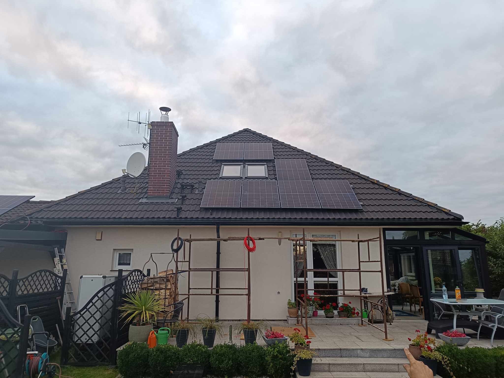 Instalacja paneli słonecznych na dachu domu jednorodzinnego, widoczne rusztowania, komin z anteną satelitarną, i fragment ogrodu z tarasem.
