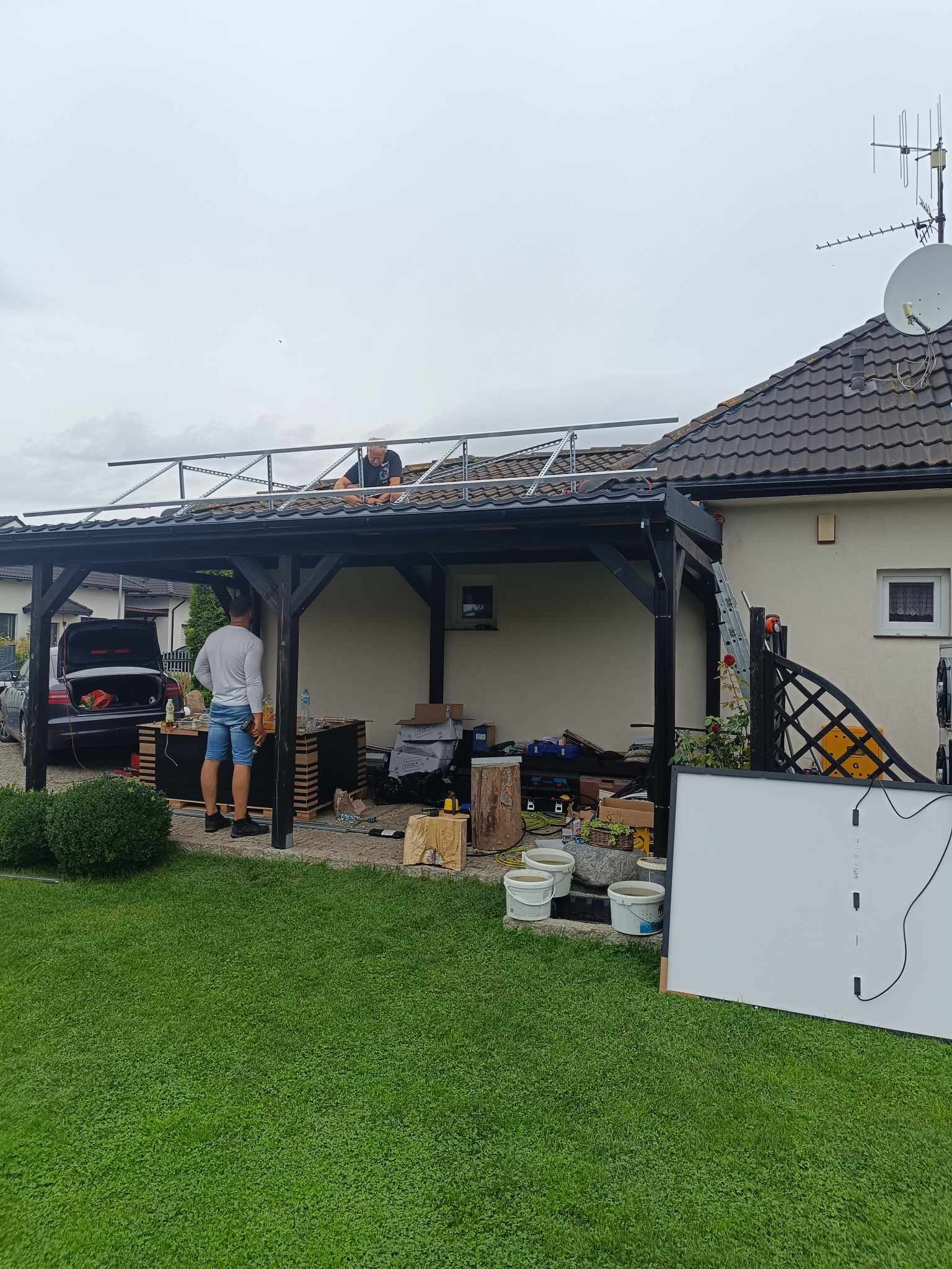 Montaż metalowej konstrukcji pod panele słoneczne na dachu wiaty garażowej, dwóch monterów przy pracy, narzędzia i skrzynki porozstawiane wokół, trawnik przed domem.