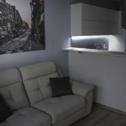 Jasnoszara ściana z obrazem ulicy, biała skórzana sofa z ciemnymi poduszkami i fragment zabudowy kuchennej z oświetleniem LED pod szafkami.