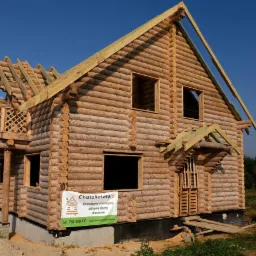 Surowy dwukondygnacyjny dom z bali w trakcie budowy, widoczne niezabudowane otwory okienne, drewniana konstrukcja dachu i balkon z ozdobną balustradą, na pierwszym planie tablica reklamowa firmy.