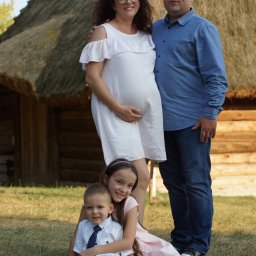 FOTO WERDMAN sp. z o.o. - Uśmiechnięta, ciężarna kobieta w białej sukience z mężem i dwójką dzieci na tle chaty krytej strzechą. Sesja rodzinna na łonie natury, ciepłe światło.