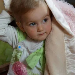 Urocze dziecko z blond włosami, otulone miękkim, beżowo-r&oacute;żowym kocykiem, w kwiecistej sukience z zieloną kokardą, spogląda w bok z wyrazem zaciekawienia.