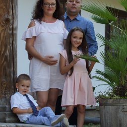 FOTO WERDMAN sp. z o.o. - Uśmiechnięta rodzina w pastelowych strojach pozuje do zdjęcia na zewnątrz. Kobieta w ciąży z mężem i dwojgiem dzieci. Chłopiec siedzi na schodach, a dziewczynka trzyma liść.