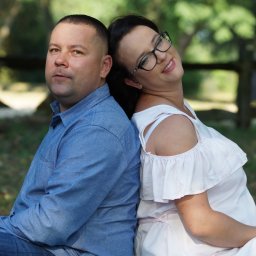 FOTO WERDMAN sp. z o.o. - Uśmiechnięta kobieta w okularach i mężczyzna w niebieskiej koszuli siedzą plecami do siebie w plenerze, na tle drzew i drewnianego płotu.