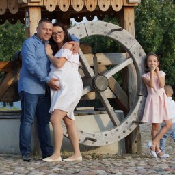 FOTO WERDMAN sp. z o.o. - Ciążowa sesja rodzinna przy drewnianej studni: uśmiechnięta para, z brzuchem w centrum uwagi, obok dzieci w pastelowych ubraniach na tle zieleni.