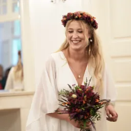 Uśmiechnięta blondynka w białej sukni i wianku z kwiatów, trzyma bukiet. W tle lustro, świecznik i fragment białych drzwi.