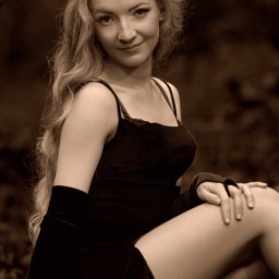 FOTO WERDMAN sp. z o.o. - Portret kobiety o długich blond włosach, ubranej w czarną sukienkę i rękawiczki, siedzącej na tle ciemnego, naturalnego otoczenia w sepiowej tonacji.