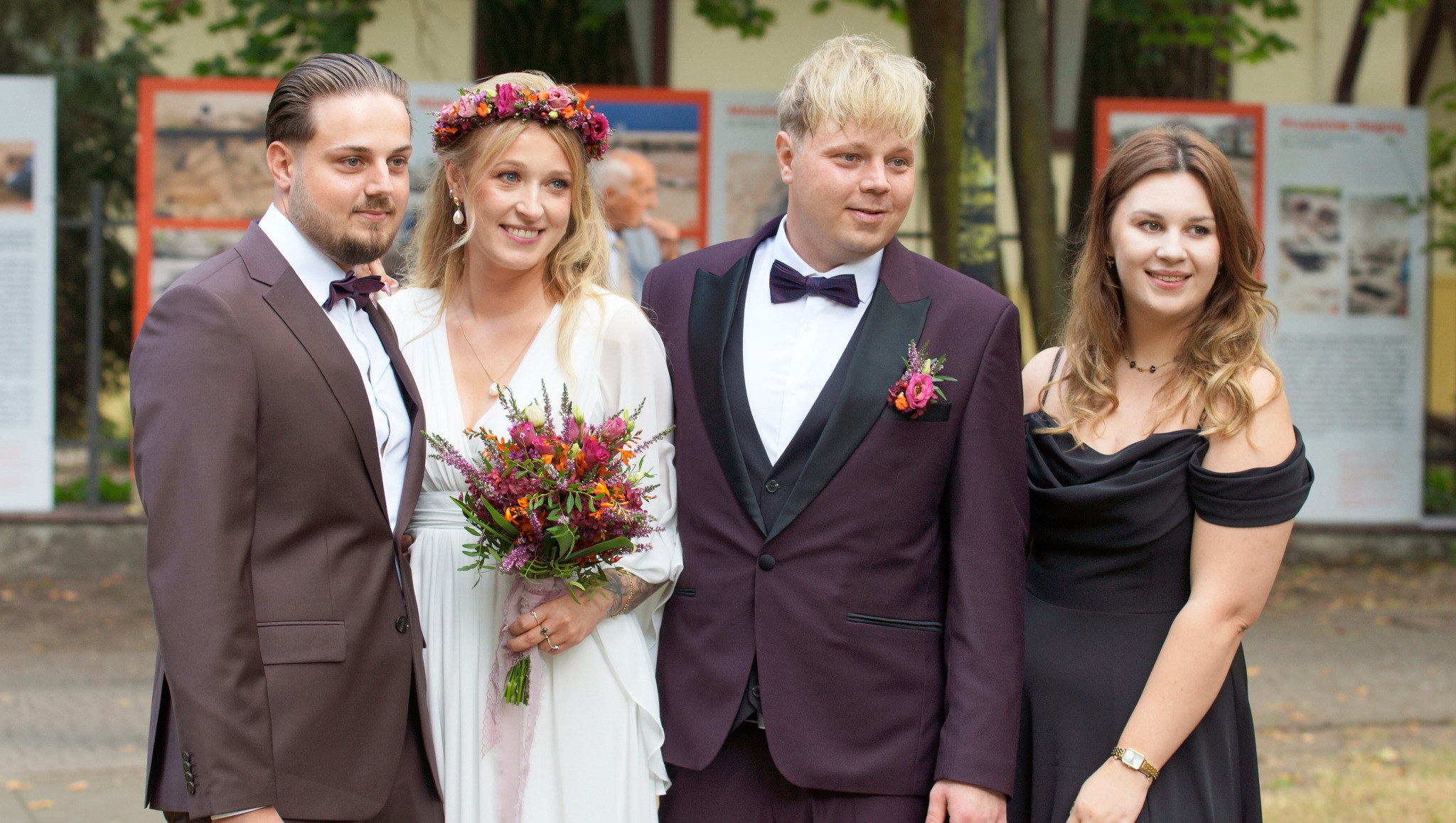 Uśmiechnięta para młoda z bukietem i goście weselni w eleganckich strojach pozują na tle tablic informacyjnych. Uwagę przykuwa wianek panny młodej i bordowy garnitur pana młodego.
