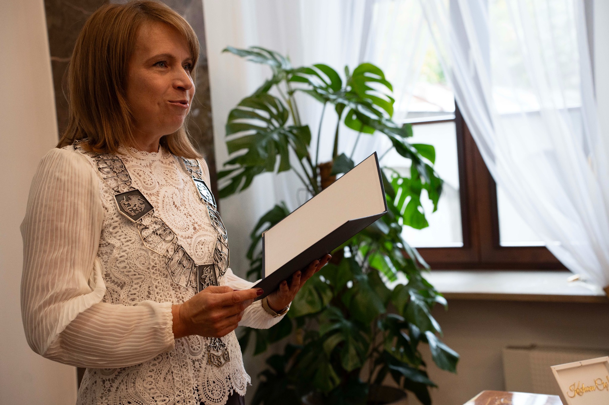Kobieta w stroju galowym z łańcuchem, trzymająca czarną teczkę na tle rośliny doniczkowej i okna z firanką. Ujęcie z profilu, jasne światło.
