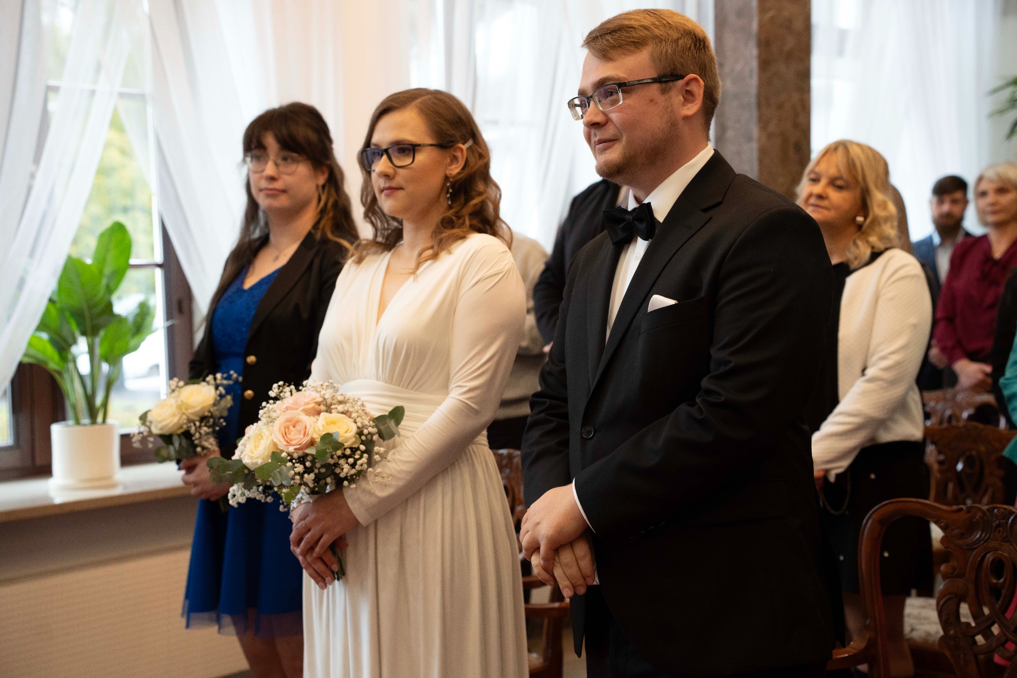 Elegancka para młoda w trakcie ceremonii ślubnej, panna młoda w białej sukni trzyma bukiet, pan młody w czarnym smokingu, w tle goście i okno.