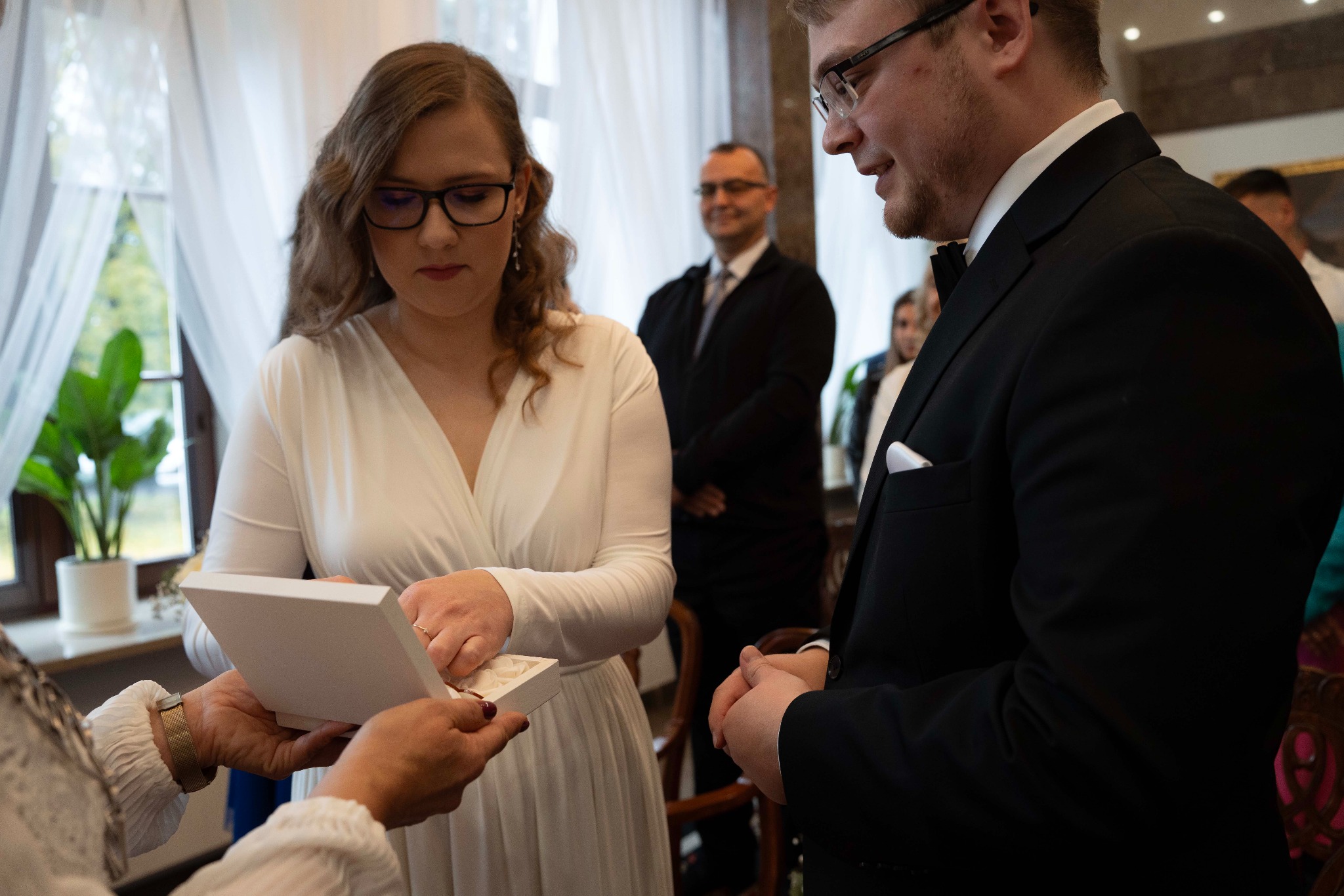 Moment wręczania obrączek podczas ceremonii ślubnej. Panna młoda w białej sukni, pan młody w czarnym garniturze. W tle goście i okno z rośliną doniczkową.