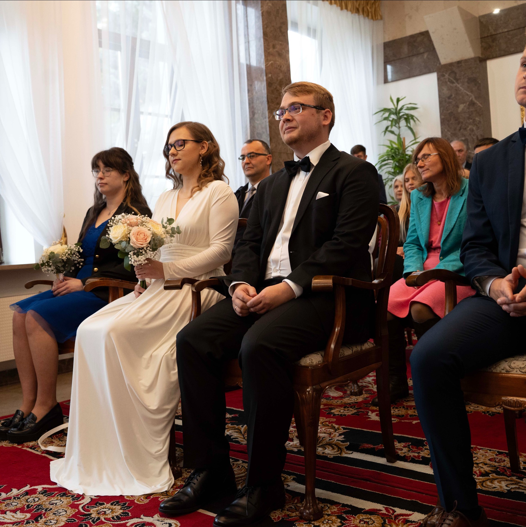 Para młoda w trakcie ceremonii ślubnej siedzi na krzesłach, trzymając bukiety. W tle goście i dekoracje sali. Ujęcie z boku, naturalne światło.