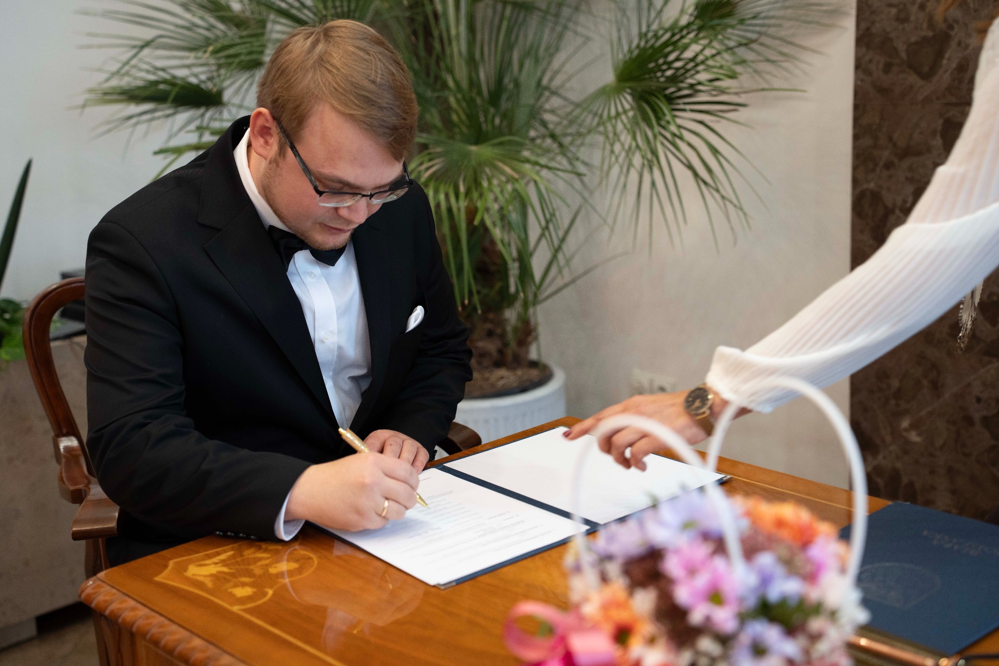 Pan młody w czarnym smokingu podpisuje dokument na drewnianym stole. W tle roślina doniczkowa i fragment kobiety w białej bluzce. Bukiet kwiatów w rogu kadru.