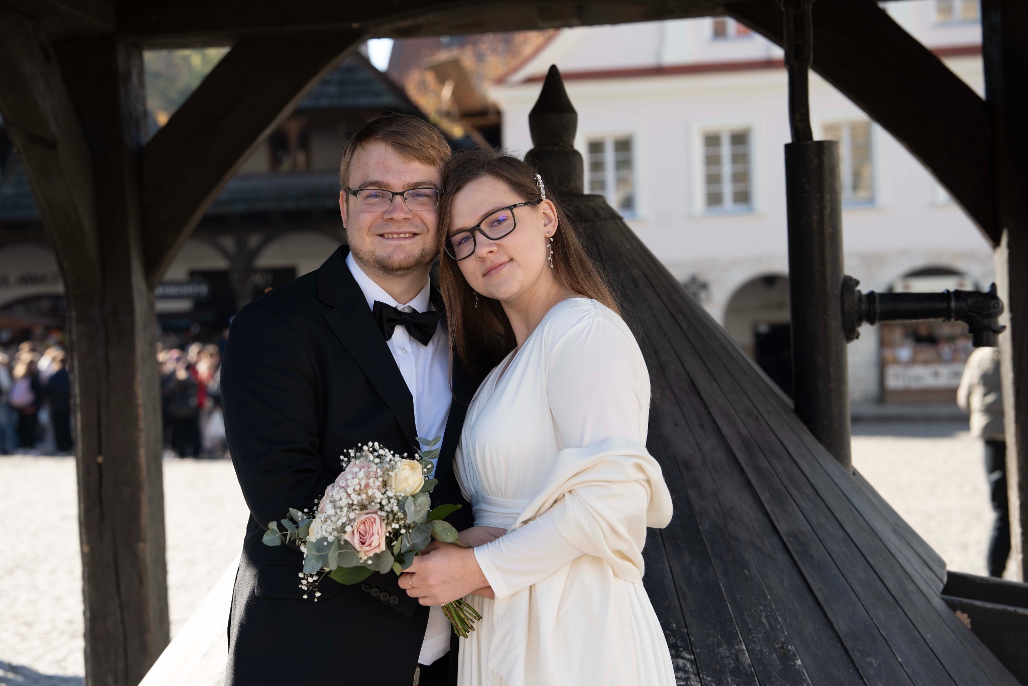 Uśmiechnięta para młoda w okularach, pozująca z bukietem na tle drewnianej konstrukcji i jasnego budynku w słoneczny dzień. Elegancki strój, naturalne światło.