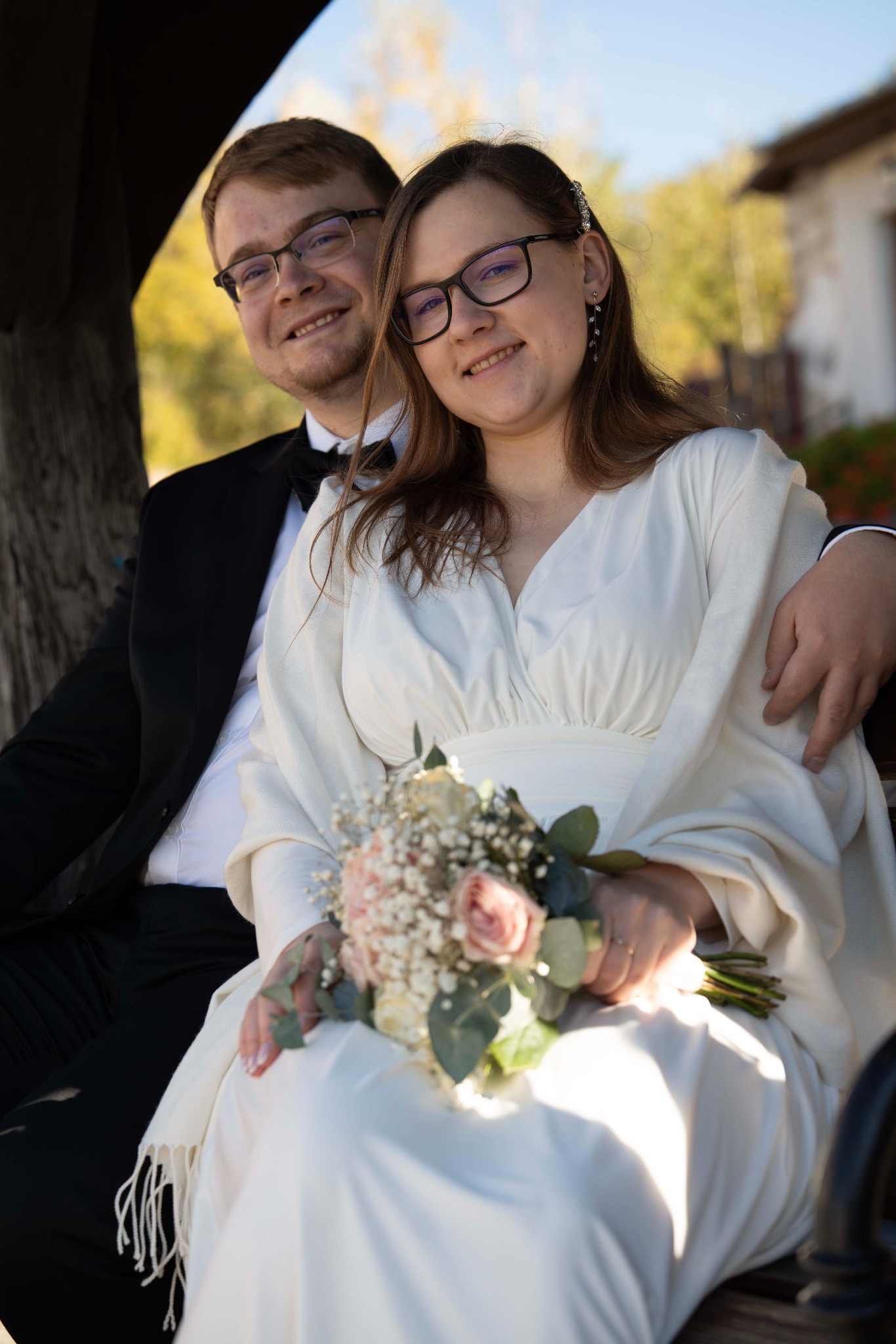 Uśmiechnięta para młoda w okularach siedzi na ławce. Panna młoda trzyma bukiet z różowych i białych kwiatów. Pan młody w czarnym smokingu z muszką.