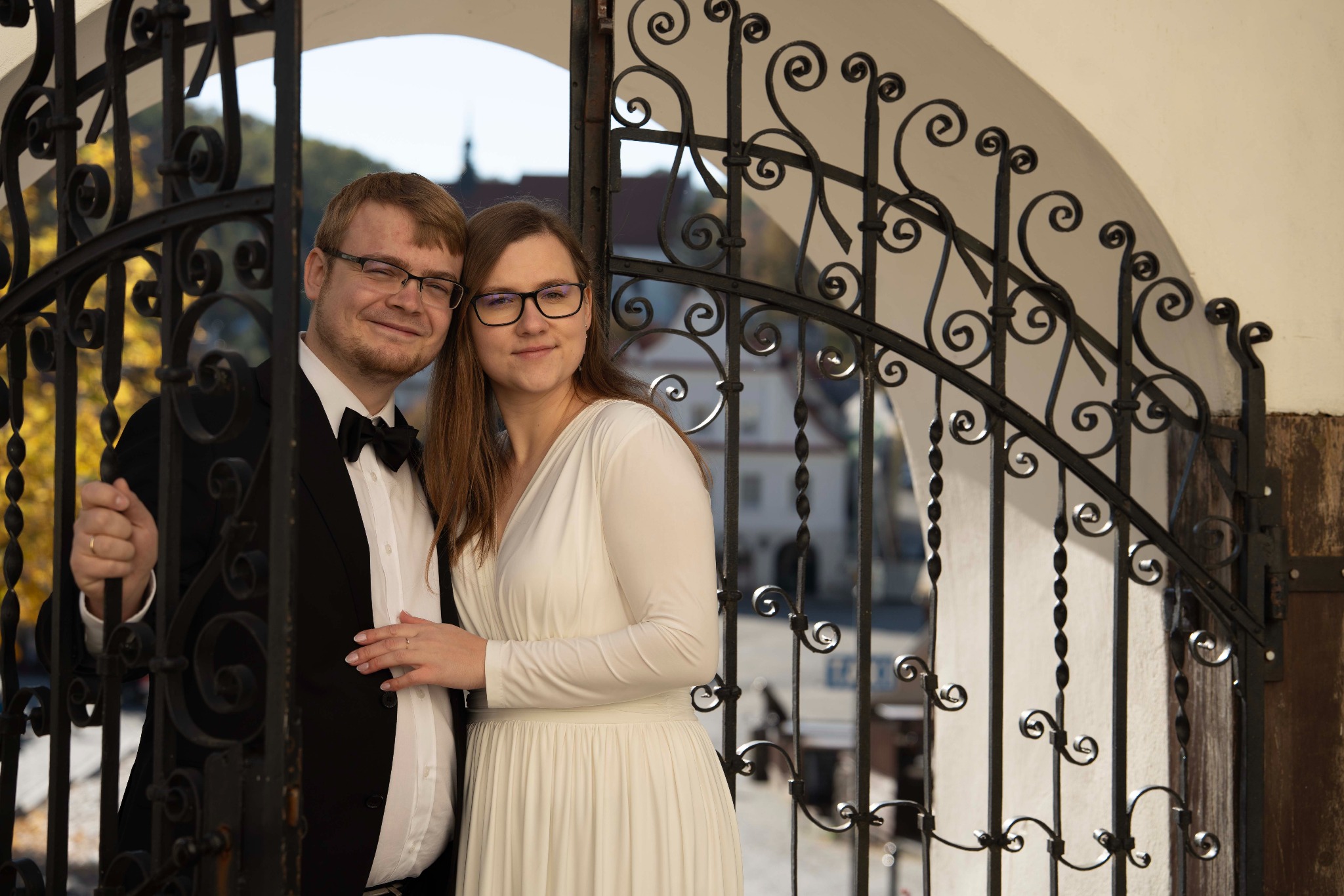 Uśmiechnięta para młoda w okularach, pozuje za ozdobną, metalową bramą na tle zabudowań miejskich. Pan w smokingu, pani w białej sukni.