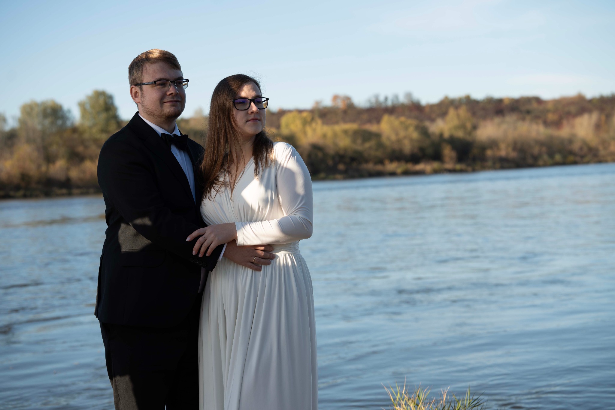 Para w strojach ślubnych pozuje na tle rzeki i jesiennego krajobrazu. Pan w czarnym smokingu, pani w białej sukni. Ujęcie w naturalnym świetle, z delikatnym rozmyciem tła.