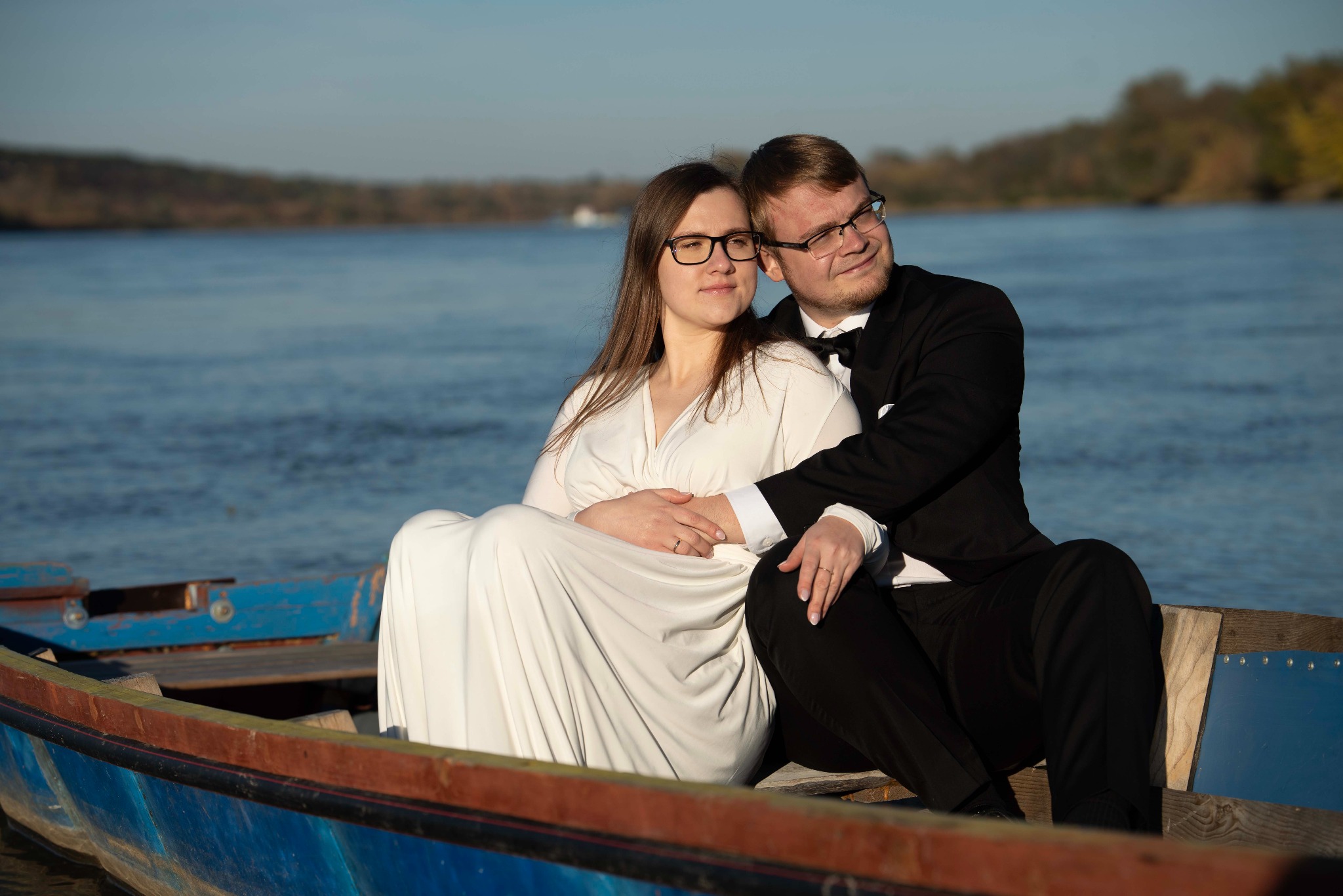 Zakochana para w łodzi na tle wody i drzew. Kobieta w białej sukience, mężczyzna w garniturze z muszką. Romantyczna sesja plenerowa w słoneczny dzień.
