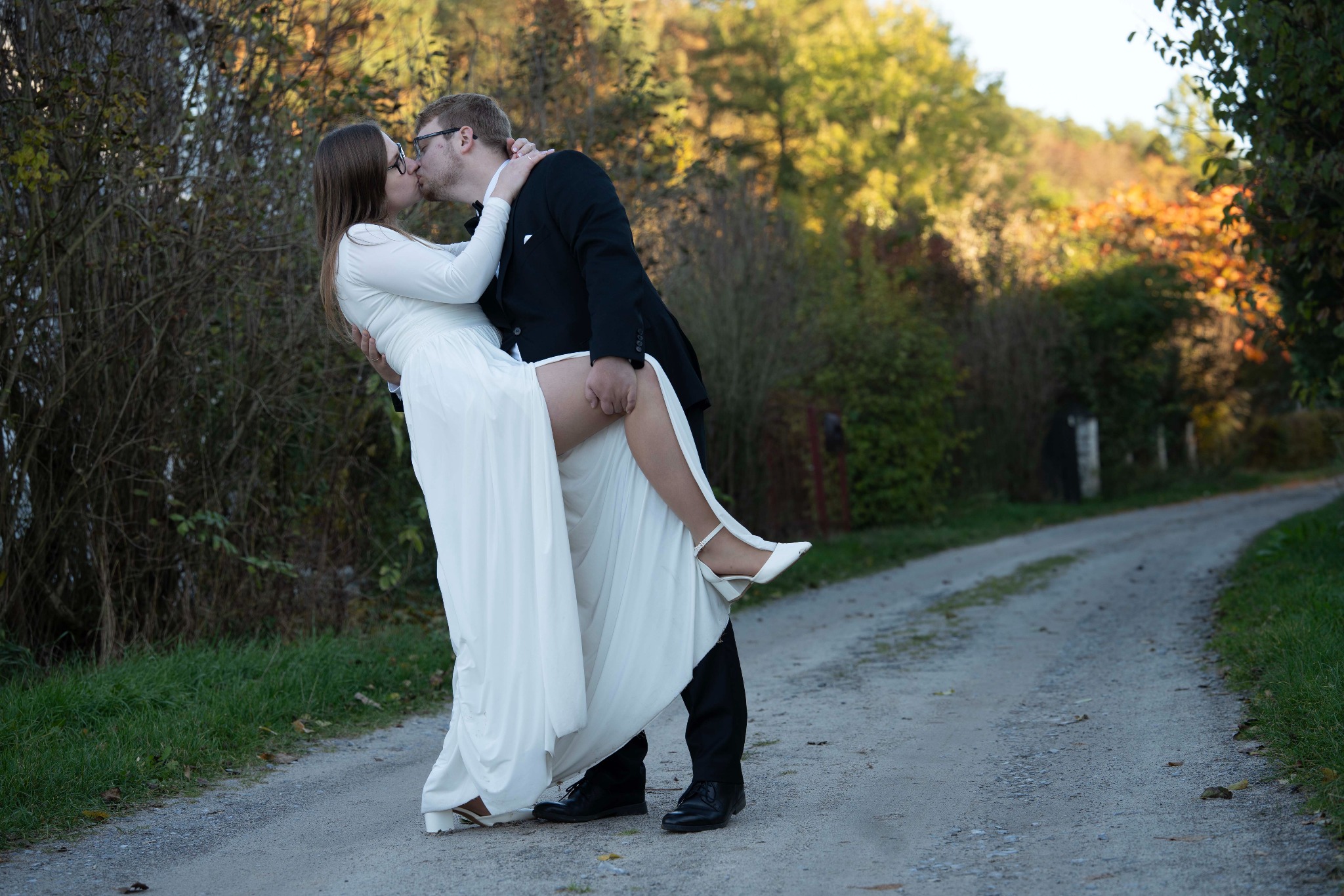 Para młoda w plenerze: Panna Młoda w białej sukni, unoszona przez Pana Młodego w czarnym garniturze, całuje się na leśnej drodze. Romantyczna sesja ślubna w naturalnym otoczeniu.