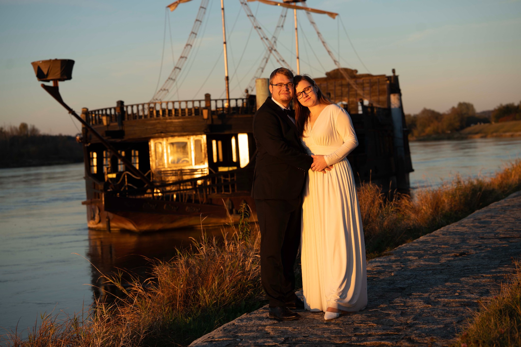 Para w eleganckich strojach pozuje na tle rzeki i stylizowanego statku w ciepłym, zachodzącym słońcu. Romantyczna sceneria i stonowana kolorystyka.