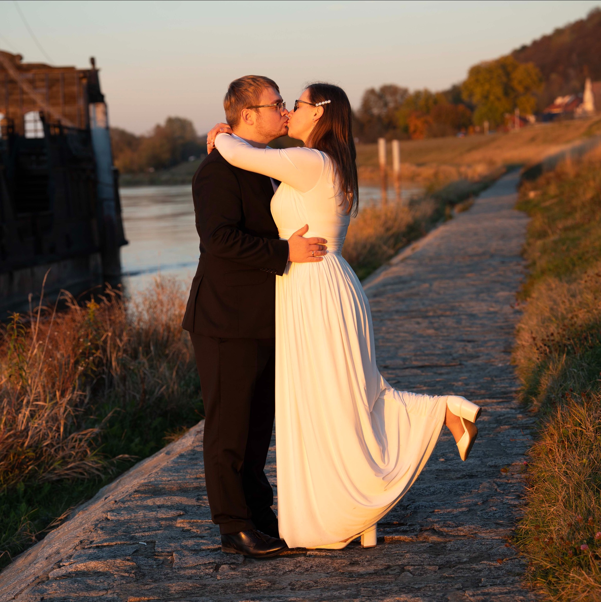 Pocałunek pary młodej w plenerze, ona w białej sukni i butach na obcasie, on w czarnym garniturze, w tle rzeka i zieleń w ciepłym świetle zachodzącego słońca.