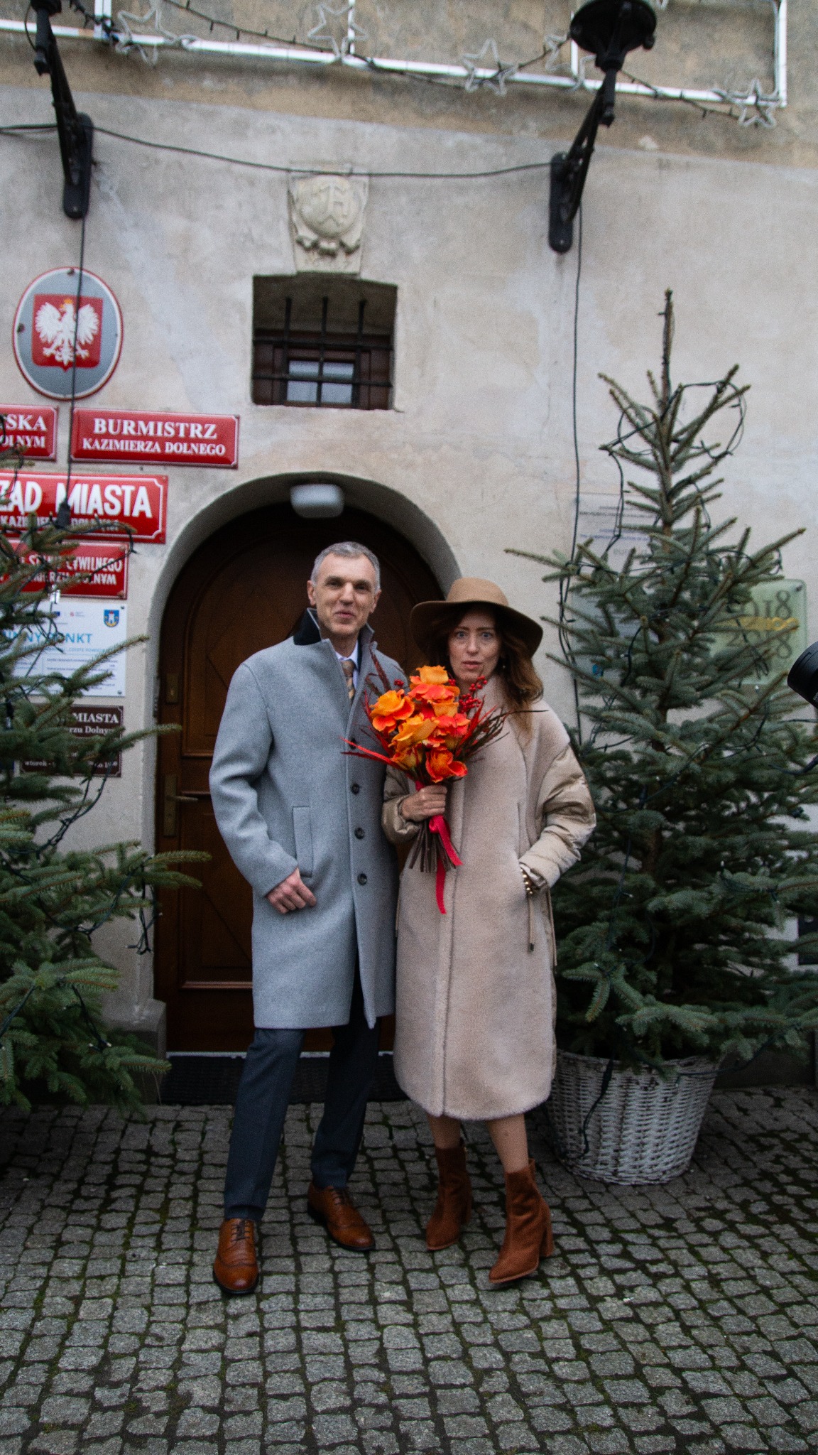 Elegancka para w Kazimierzu Dolnym: mężczyzna w szarym płaszczu, kobieta z bukietem w beżowym płaszczu i kapeluszu, na tle kamienicy z tabliczkami urzędowymi.
