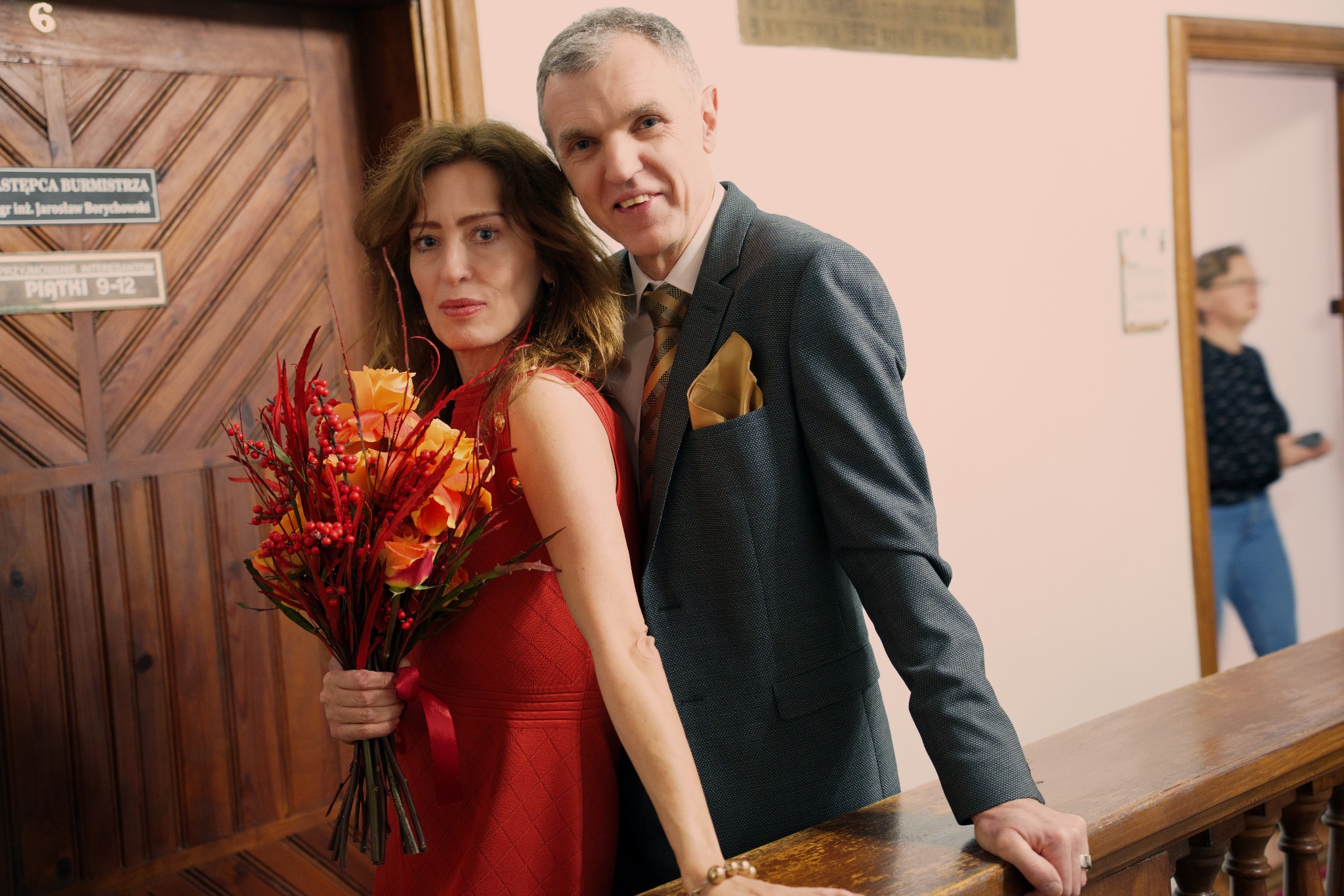 Elegancka para pozuje z bukietem pomarańczowych róż na tle drewnianych drzwi z tabliczką informacyjną w budynku urzędu. Kobieta w czerwonej sukience, mężczyzna w garniturze.