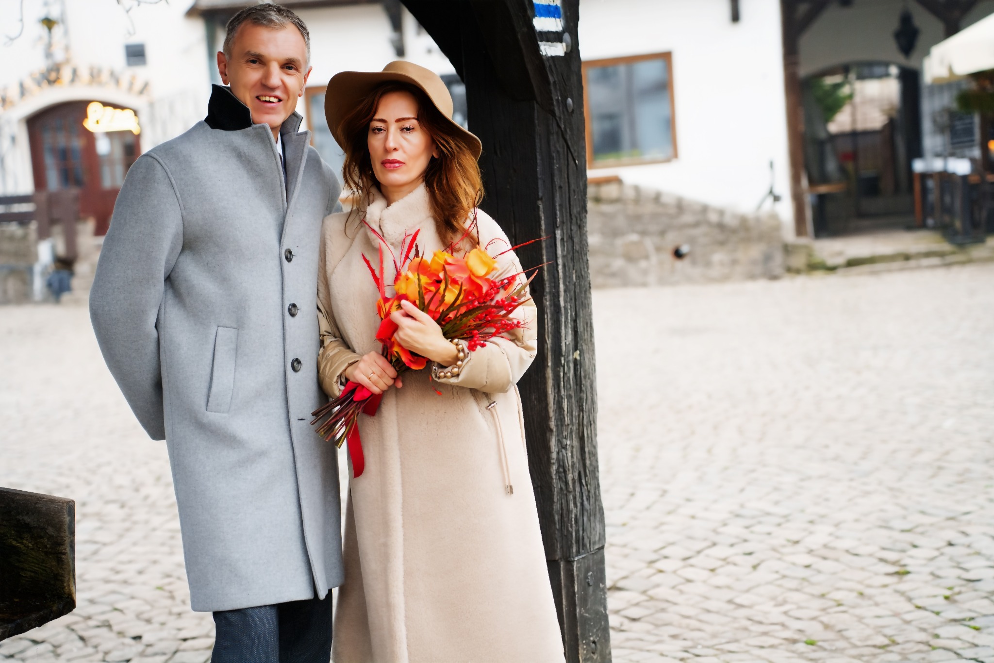 Elegancka para w jesiennej scenerii: mężczyzna w szarym płaszczu, kobieta w beżowym z bukietem, pozują na tle brukowanej uliczki i zabytkowej architektury.