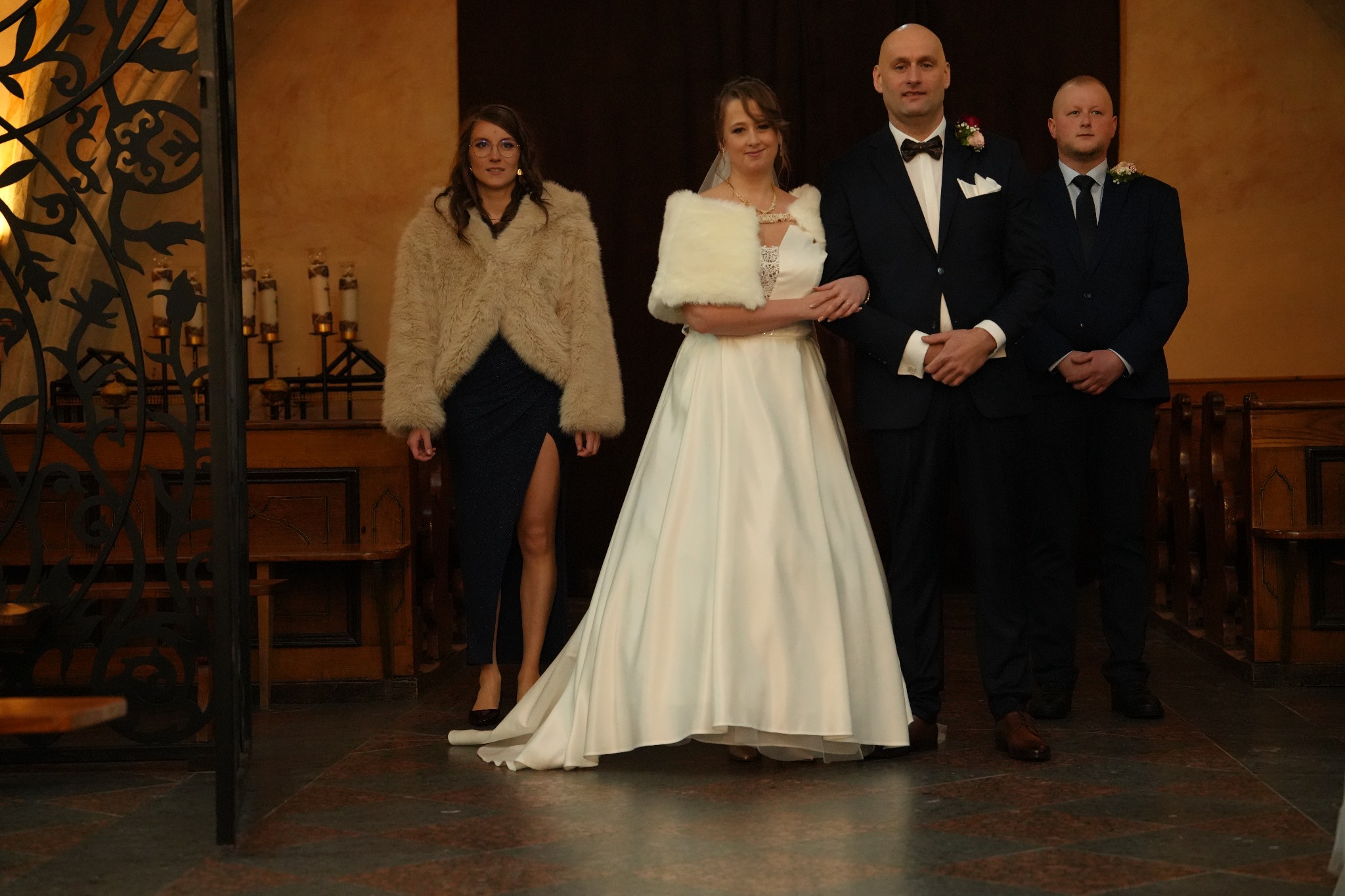 Panna młoda w białej sukni i futrzanym etolu, obok druhny w futrze i rozciętej sukni, oraz pan młody i drużba w ciemnych garniturach, stoją w kościele.