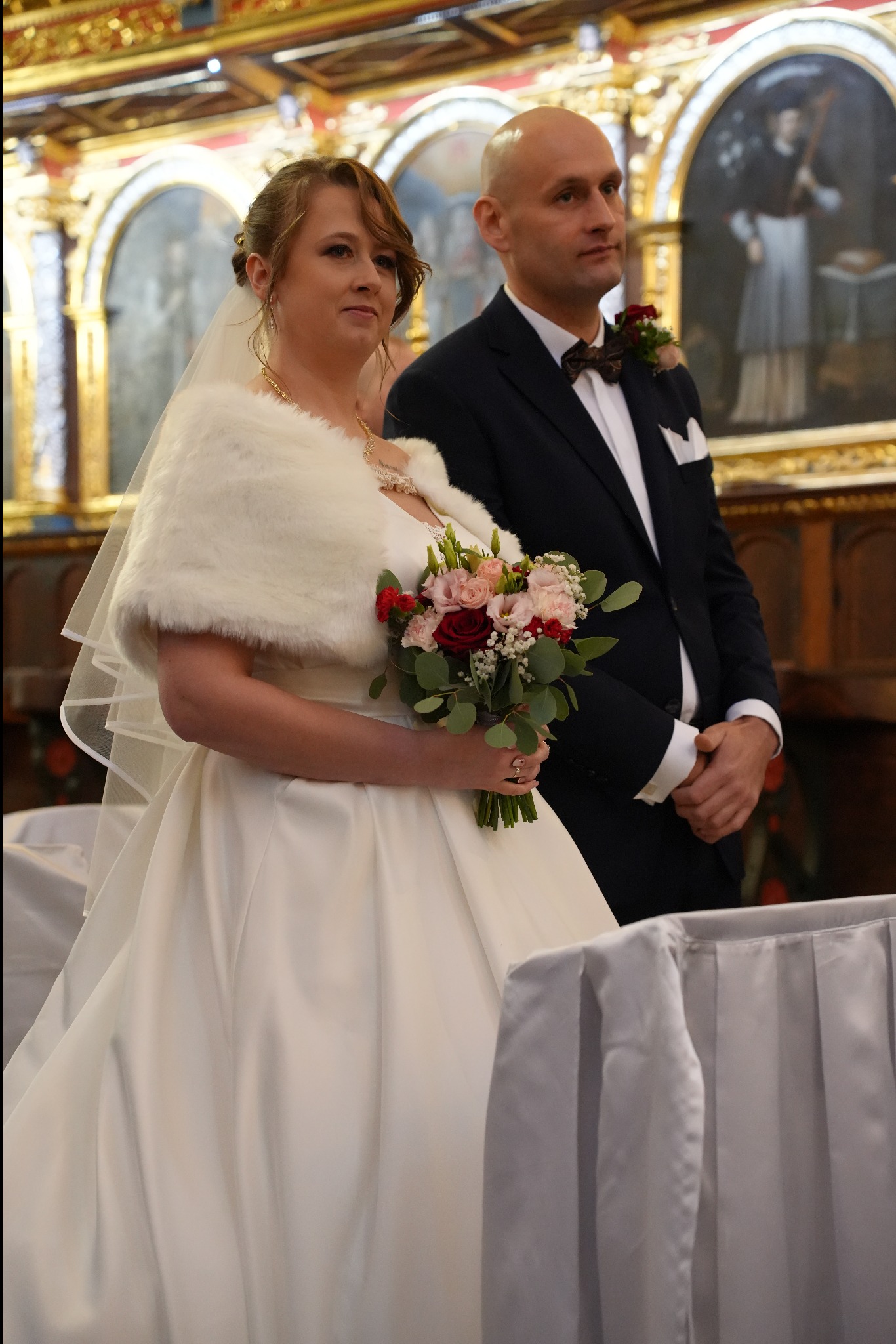 Para młoda w kościele: panna młoda w białej sukni z futrzanym szalem trzyma bukiet, obok pan młody w granatowym garniturze z muszką. W tle obrazy i złocenia.
