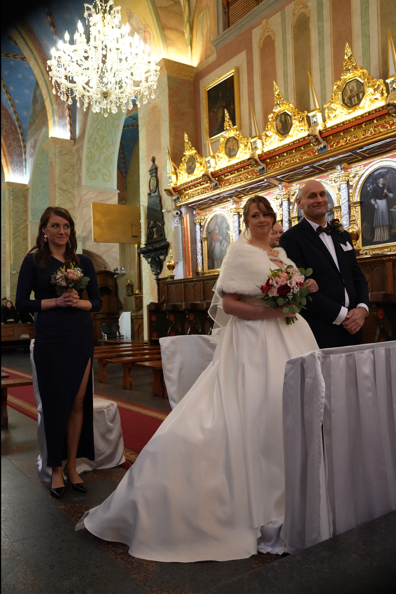 Para młoda w kościele podczas ceremonii ślubnej, panna młoda w białej sukni z futrzanym szalem, pan młody w czarnym smokingu, druhna z bukietem kwiatów.