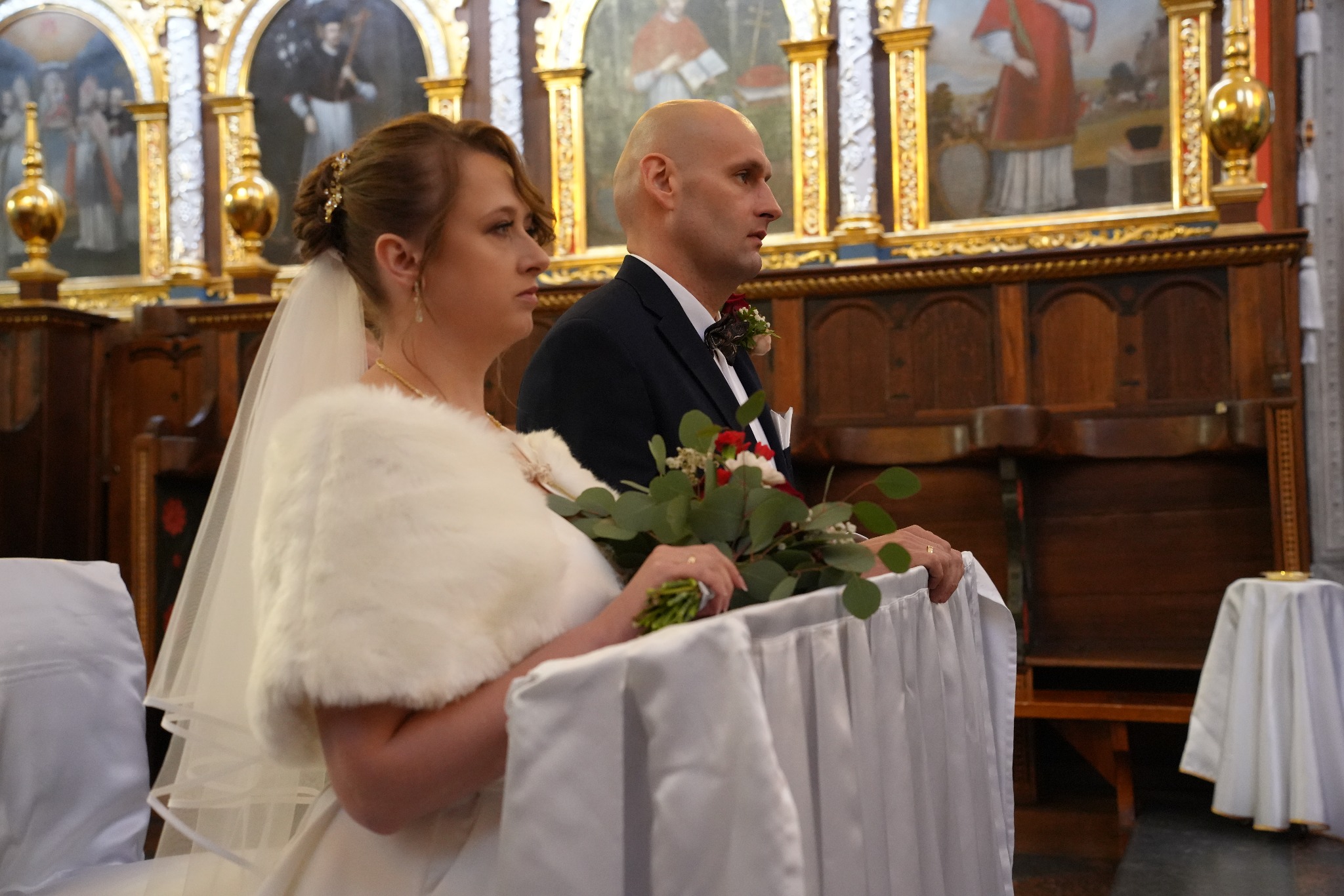 Para młoda w kościele w Łęcznej podczas ceremonii ślubnej. Panna młoda w futerku trzyma bukiet, a pan młody w garniturze stoi obok. W tle zabytkowe obrazy.