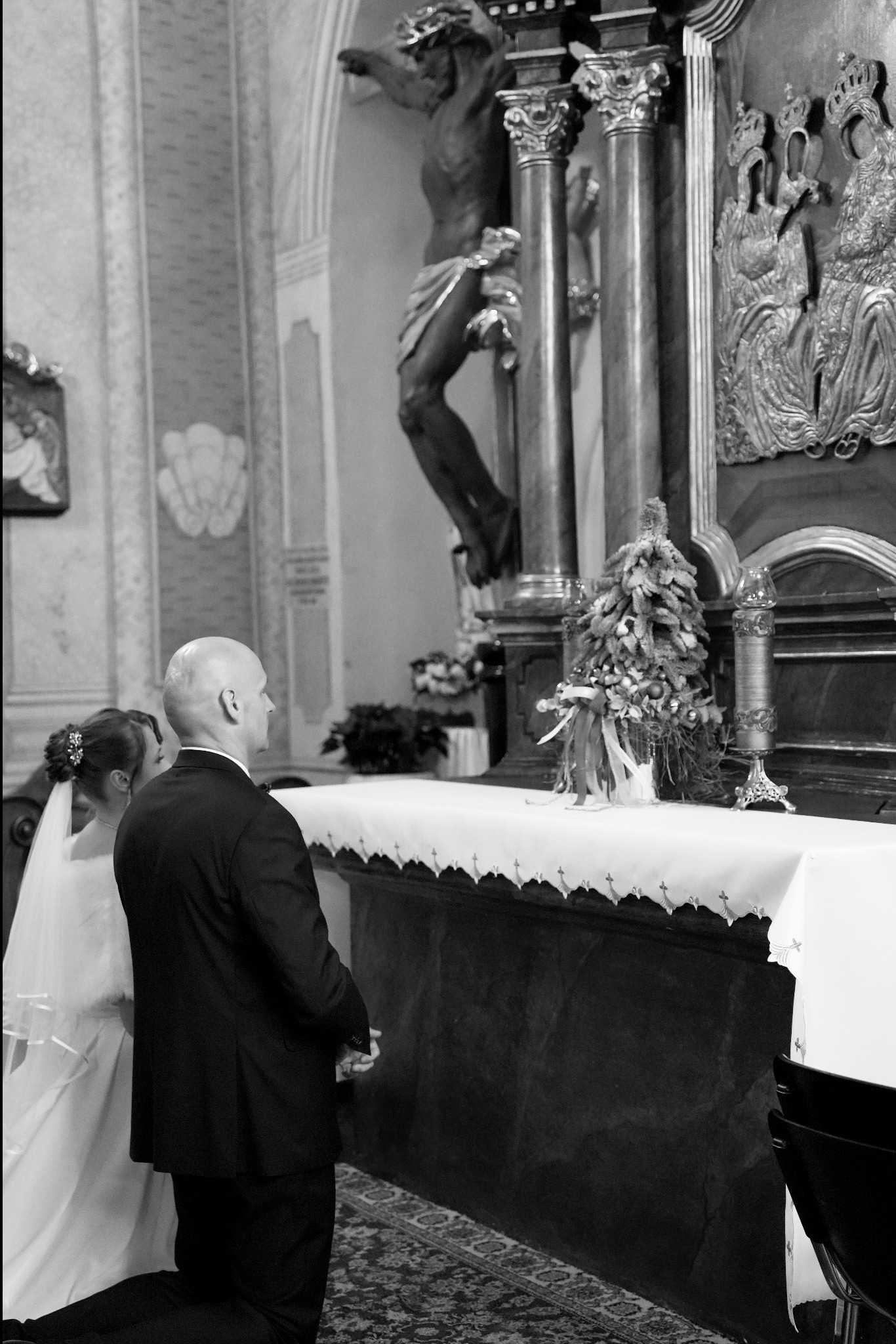 Czarno-białe zdjęcie pary młodej klęczącej przed ołtarzem w kościele, z widocznym krucyfiksem i dekoracjami. Pan młody w ciemnym garniturze, panna młoda w welonie.