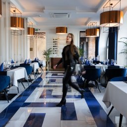 Katarzyna Gruca PROJEKTOWANIE - Eleganckie wnętrze restauracji z marmurową podłogą w biało-niebieskie pasy, okrągłymi miedzianymi lampami i stolikami nakrytymi białymi obrusami z granatowymi bieżnikami i niebieskimi serwetkami,...