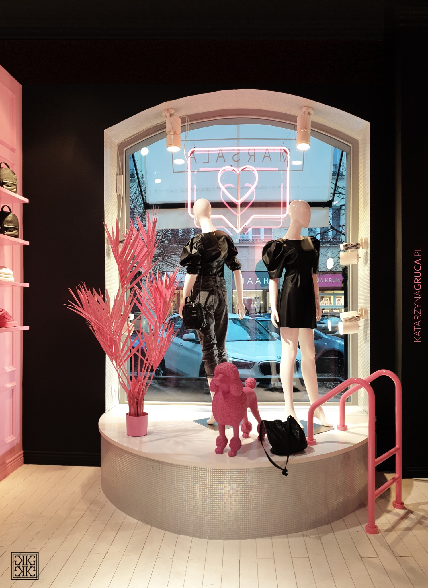 Wystawa sklepowa z dwoma manekinami w czarnych strojach, neonowym napisem MARSAR i różowym pudlem na podeście z mozaiki.