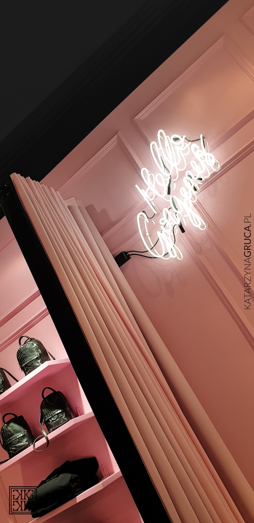 Różowe wnętrze butiku z neonowym napisem 'Hello Gorgeous', półki z czarnymi plecakami ze skóry krokodyla i zasłona w prążki.