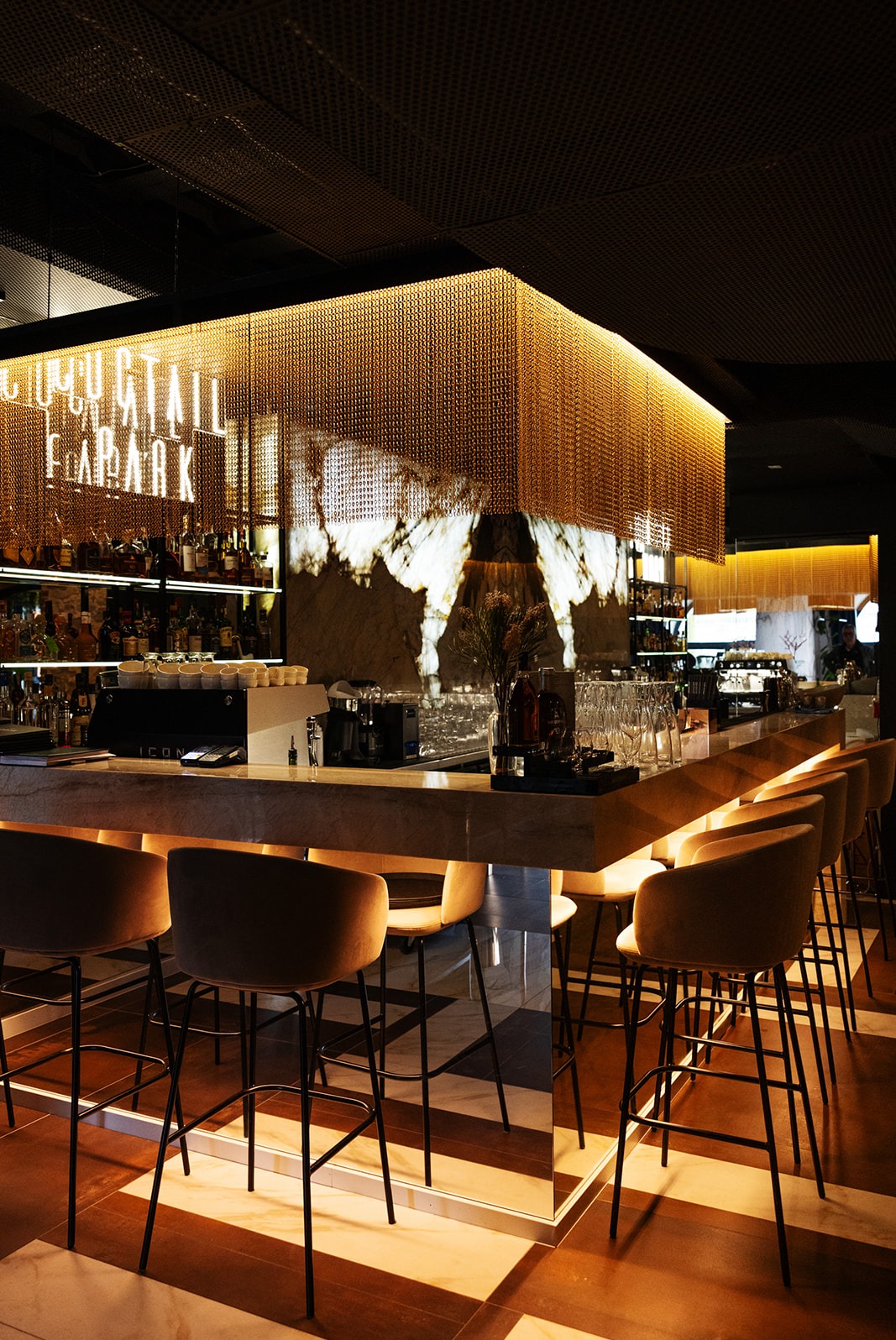 Nowoczesny bar z marmurowym blatem, złotą metalową siatką nad barem i neonowym napisem 'COCKTAIL BAR PARK', oświetlony ciepłym światłem, z rzędem krzeseł barowych z brązowymi siedziskami.