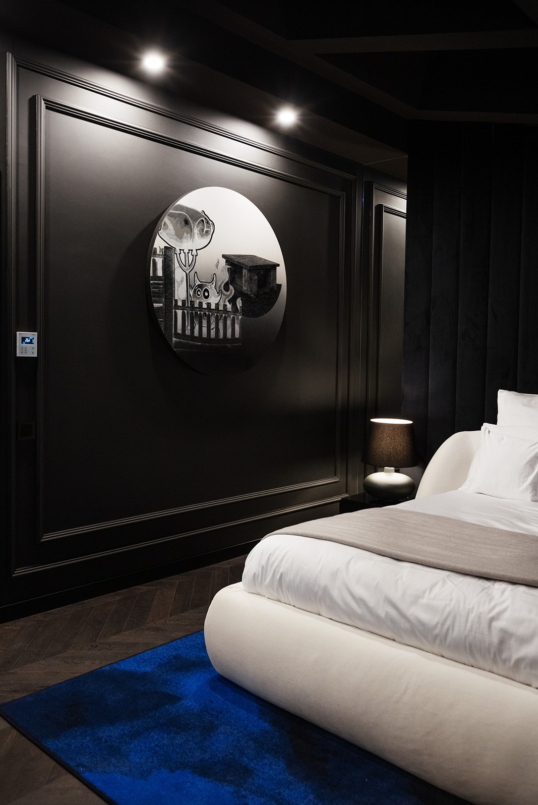 Elegancka sypialnia w ciemnej tonacji z okrągłym lustrem z abstrakcyjnym motywem, białe łóżko z beżową narzutą i niebieski dywan, oświetlenie punktowe.