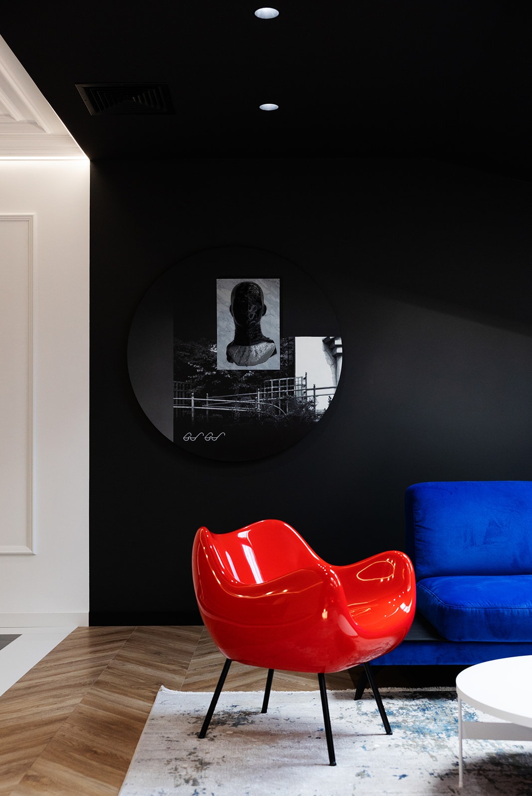 Nowoczesne wnętrze z czarną ścianą, okrągłym lustrem z kolażem, czerwoną, designerską krzesłem i niebieską sofą.