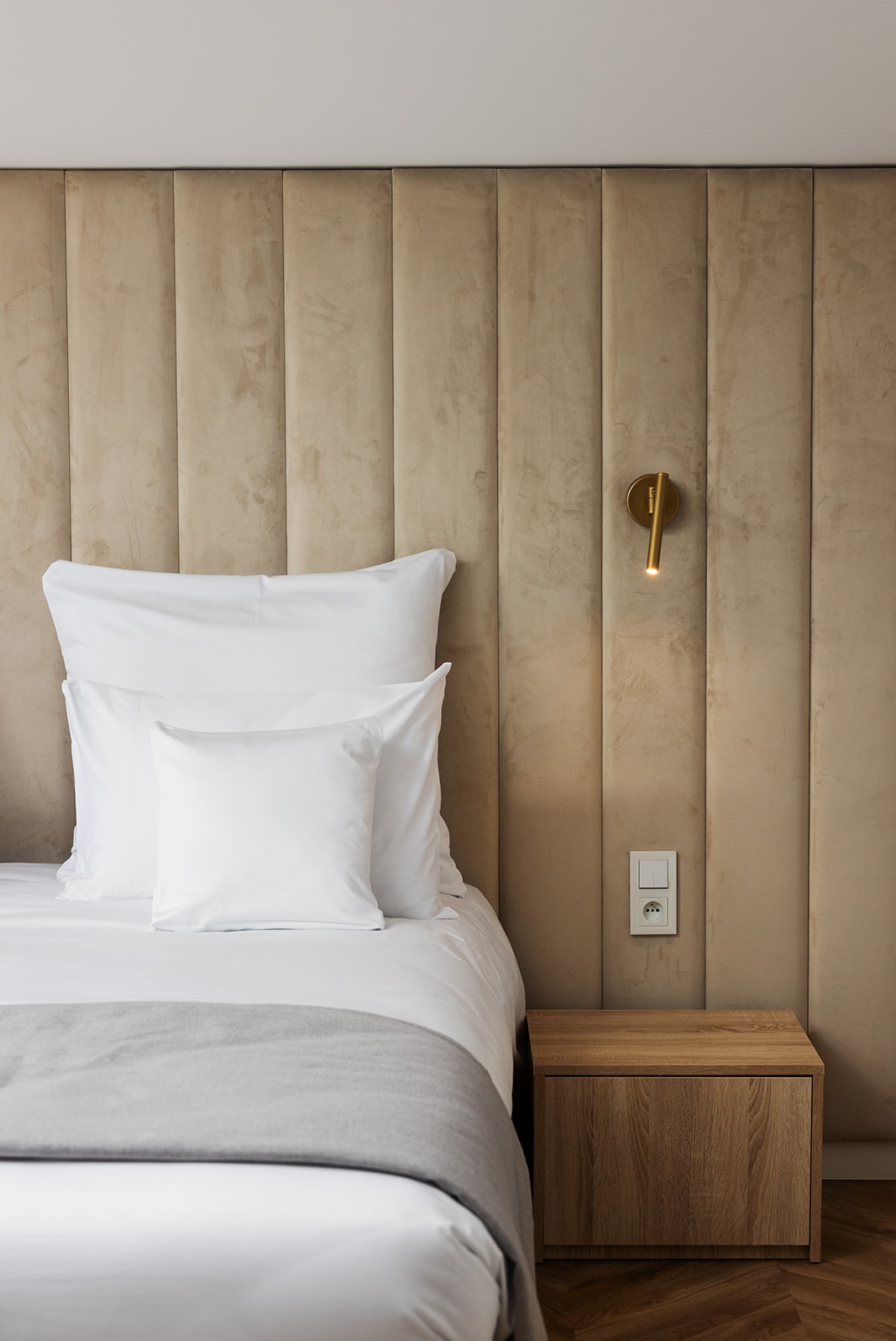 Nowoczesna sypialnia z beżowym panelem tapicerowanym za łóżkiem, białą pościelą, szarą narzutą, drewnianą szafką nocną i złotą lampką ścienną