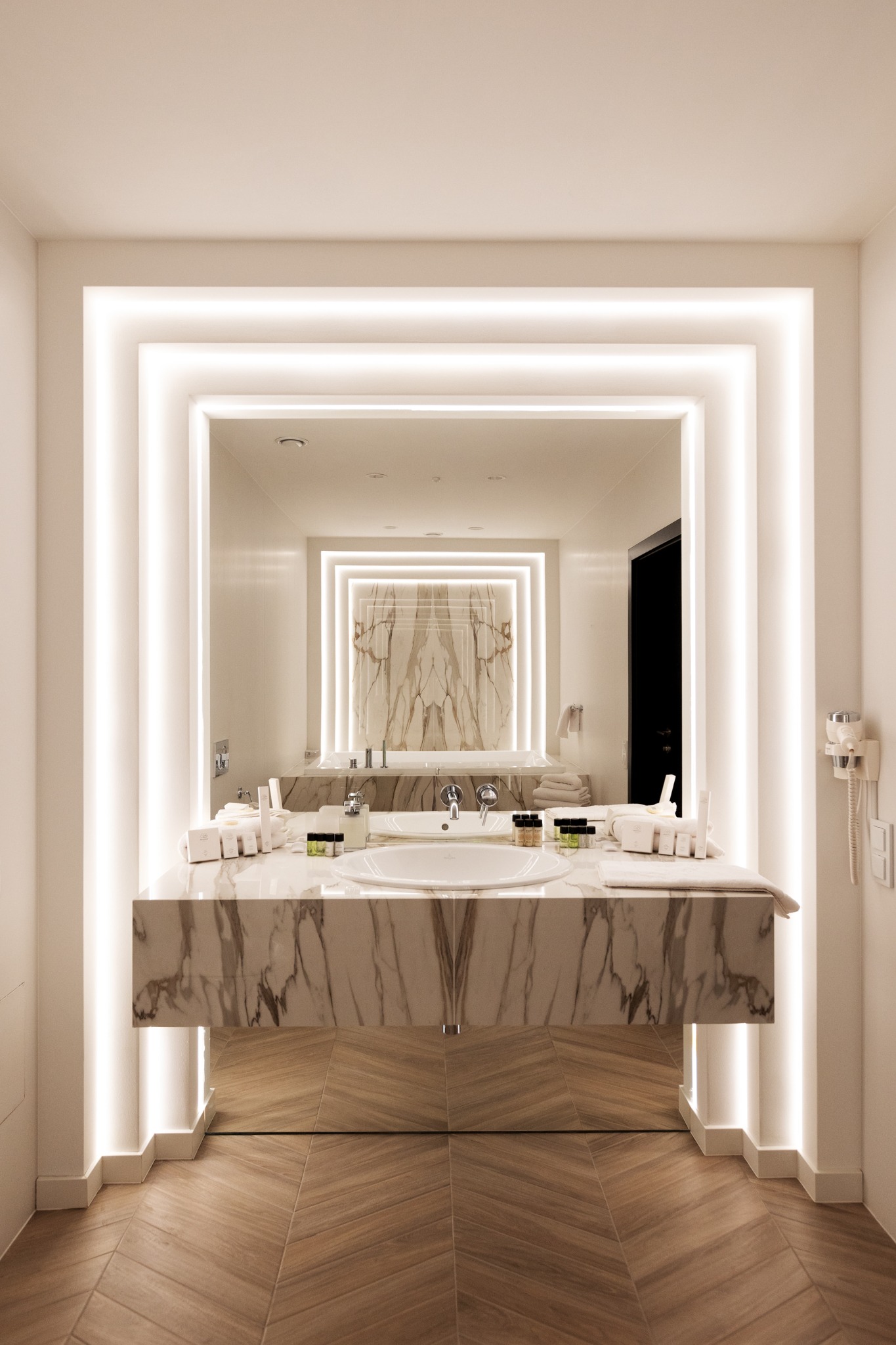 Elegancka łazienka z marmurowym blatem umywalki, podświetlanym lustrem w geometrycznej ramie i drewnianą podłogą ułożoną we wzór jodełki.