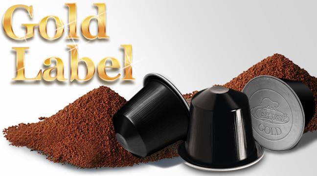 Trzy czarne kapsułki kawy, dwie ułożone obok siebie i jedna odwrócona, otoczone kopcami zmielonej kawy, z napisem 'Gold Label' w złotych literach w tle.