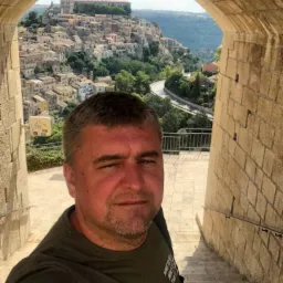 Mężczyzna robiący selfie na tle kamiennego łuku z widokiem na miasto na wzgórzu i krętą drogę.