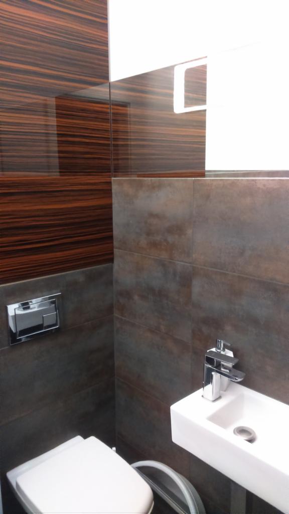 Nowoczesna łazienka z brązowymi płytkami imitującymi drewno i szarymi kaflami, biała umywalka z chromowaną baterią oraz toaleta wisząca z przyciskiem spłukującym.