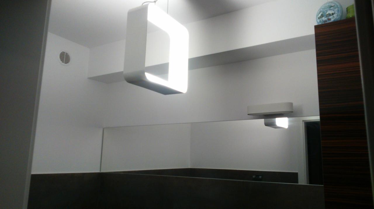 Nowoczesna łazienka z podwieszaną, kwadratową lampą LED i minimalistycznym oświetleniem nad lustrem, uzupełniona elementami dekoracyjnymi na szafce.