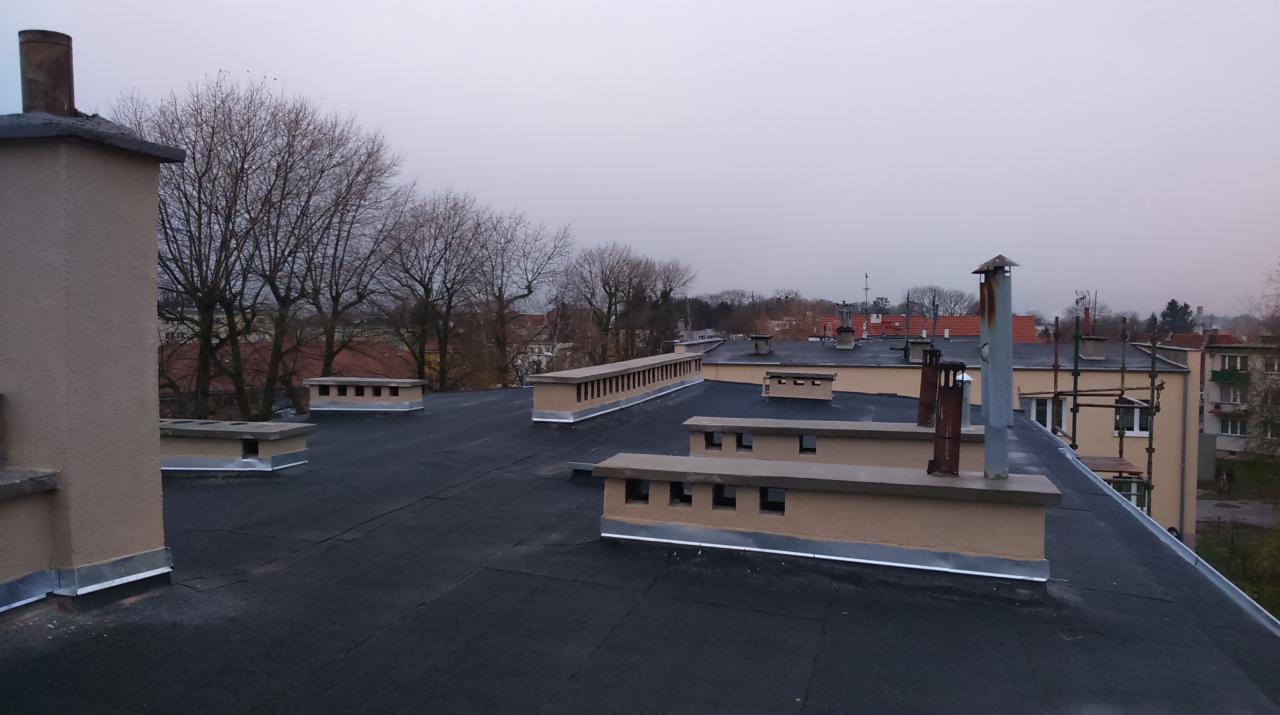 Widok na płaski dach pokryty czarną papą, z licznymi kominami wentylacyjnymi i innymi elementami infrastruktury dachowej, na tle zabudowy miejskiej w pochmurny dzień.