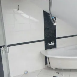 Narożna wanna na nóżkach w łazience z biało-czarnymi płytkami, widoczna kabina prysznicowa z chromowaną armaturą i fragment skośnego sufitu.