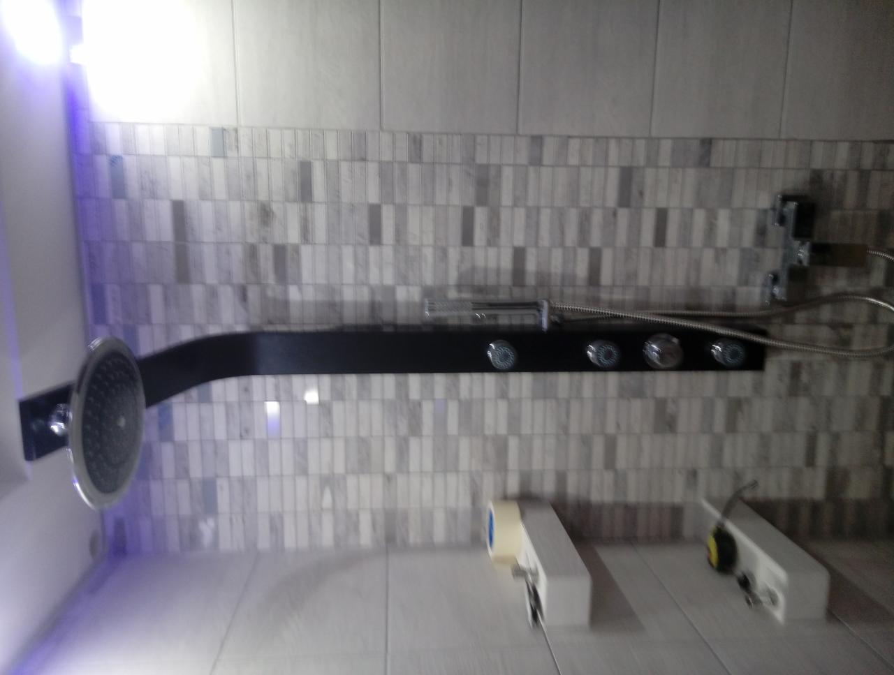 Panel prysznicowy z deszczownicą i dyszami masującymi na tle mozaiki w odcieniach szarości, widoczne elementy instalacji elektrycznej w dolnej części.