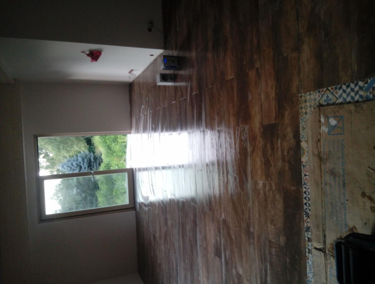 Układanie płytek imitujących drewno na ścianie w łazience, widoczne okno z widokiem na zieleń i przykryte folią zabezpieczającą podłoże.